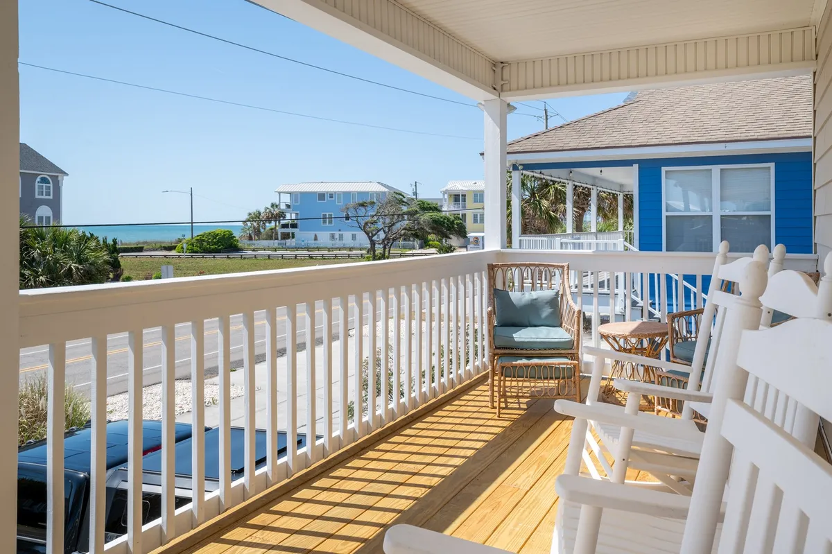 This image features a charming front porch adorned with classic rocking chairs, offering a sweeping view of the ocean. The blue house and swaying palm trees frame a tranquil outdoor sitting area perfect for enjoying coffee, reading, or simply soaking in the coastal air. The mood is peaceful and friendly, inviting relaxation and connection with the outdoors.