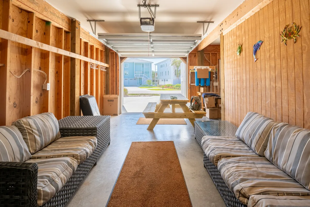 A relaxed garage-lounge space with woven sofas, a large picnic table, and an open garage door that frames the transition to the outdoors. The area is set up for socializing, shared meals, and easy access to the backyard and patios.