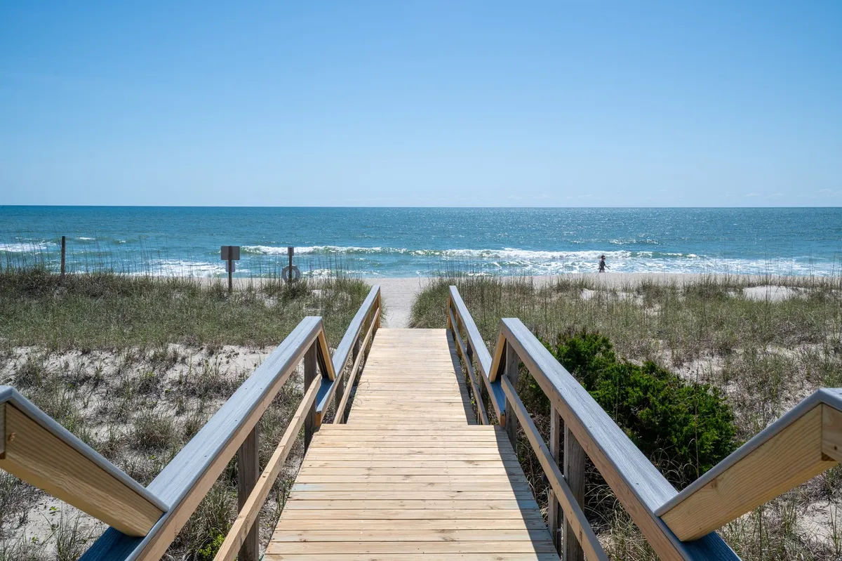 A sunlit wooden boardwalk leads from a private access point across sand dunes directly to a wide, inviting stretch of beach. The gentle waves of the ocean are visible just beyond the dunes, and a distant figure walks towards the water, evoking a sense of peaceful exploration and effortless proximity to the shoreline.