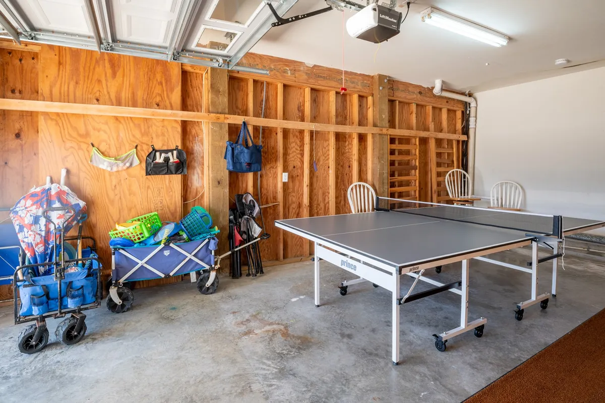 An inviting garage setup featuring a table tennis table in the foreground, flanked by open storage carts filled with sports gear. The wooden wall behind adds warmth, and there’s ample open space for movement and activity.