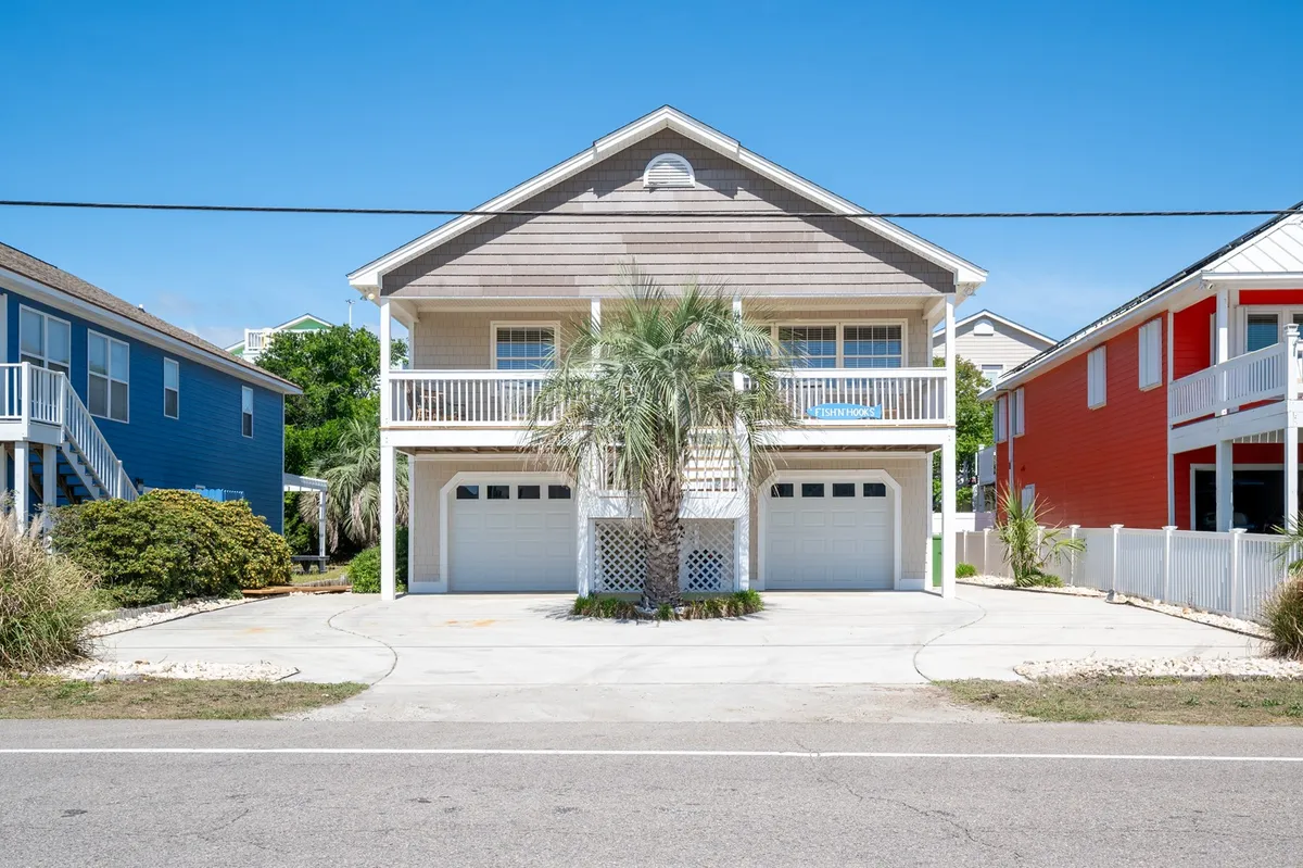 A cheerful beach house facade framed by a palm tree, with a driveway hinting at easy golf cart access. Colorful neighboring homes add to the vibrant, inviting neighborhood atmosphere, while the front porch suggests a space for sunset views and relaxed gatherings after a day by the water.