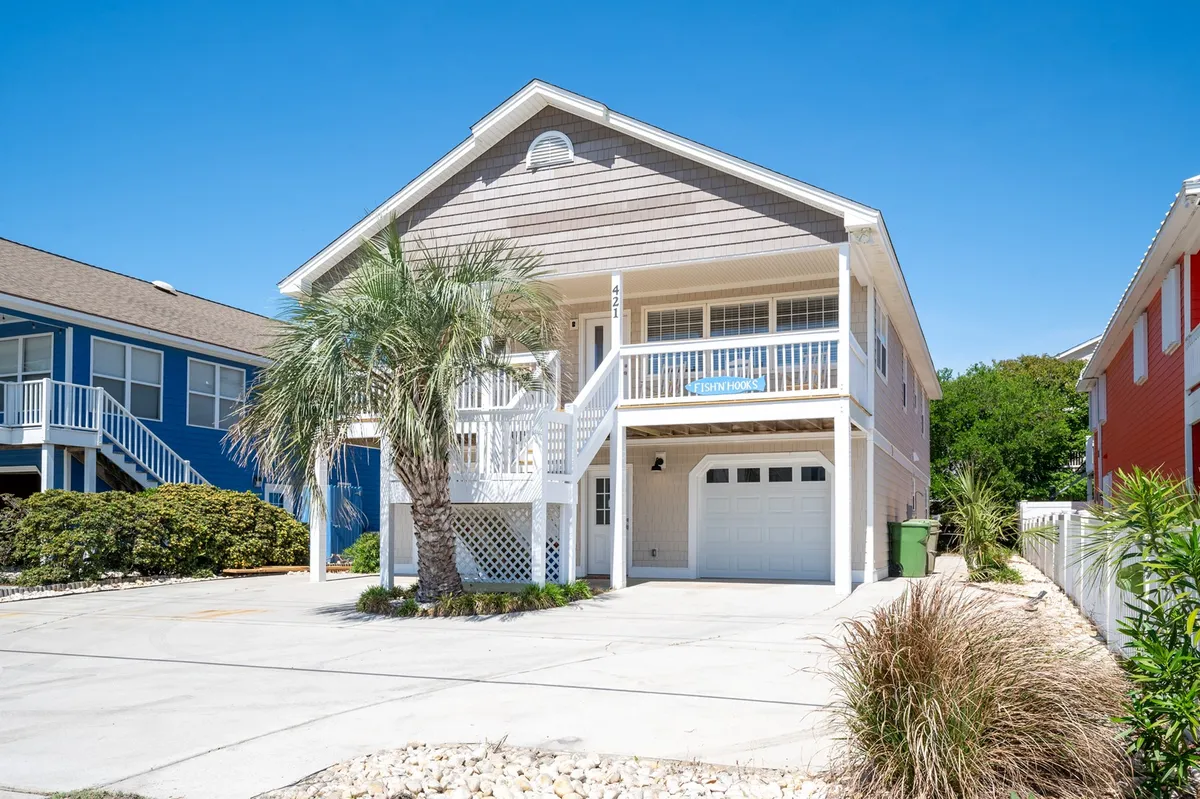 The image shows the cheerful exterior of a newly remodeled single-family beach house in Kure Beach, North Carolina. A lush palm tree stands in the foreground, framing a spacious front porch that's perfect for relaxing and watching sunsets. A distinctive blue sign reading 'Fish N Hooks' adds a playful, welcoming touch to the home's façade. The bright and inviting setting suggests a place ready for family gatherings, outdoor relaxation, and memorable summer barbecues.