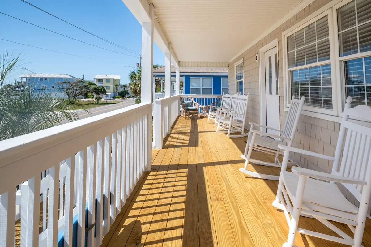 This image captures a tranquil exterior scene featuring classic rocking chairs on a wooden deck, overlooking a blue house. The setting evokes the feeling of relaxation and connection to the outdoors, with open railings and inviting seating that encourage guests to enjoy the natural surroundings.