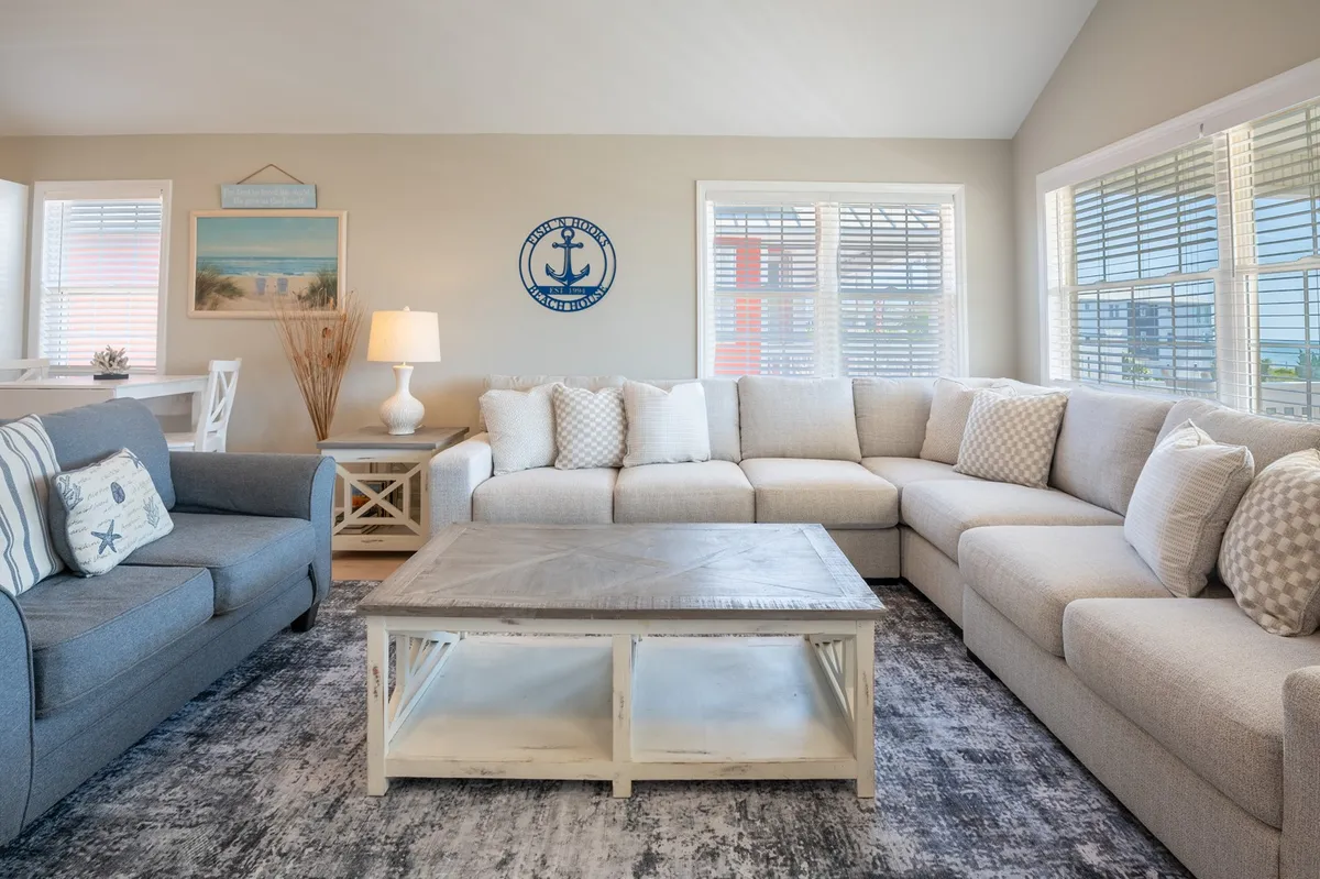 A spacious, sunlit lounge with a large sectional sofa, coffee table, nautical wall decor, and a cozy lamp. The setup invites guests to relax, play games, watch movies, or enjoy quality time with family and friends—perfectly reflecting the open-concept gathering areas described in the section.