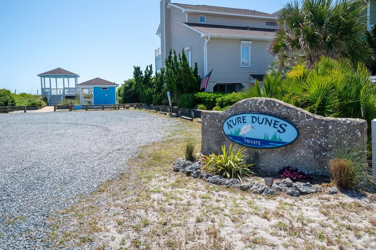 This exterior image features the welcoming Kure Dunes sign, a gravel driveway, lush greenery, and a distinctive blue gazebo. The scene evokes a sense of arrival and community, with clear connections to outdoor enjoyment and nearby beach activities.