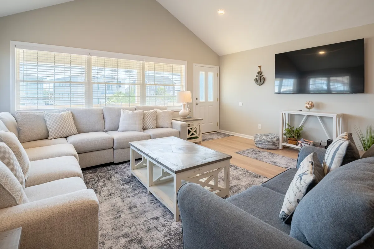 A cozy living room centered around a large sectional sofa, a coffee table, and a wall-mounted TV, all bathed in natural light from a nearby window. A lamp by the window and inviting decor make this space ideal for movie nights and casual gatherings.