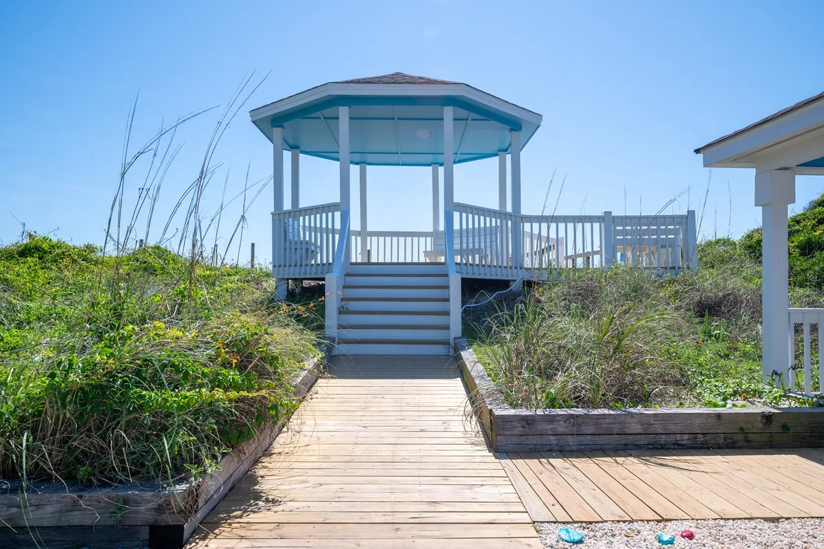 An inviting exterior scene featuring a gazebo set along a wooden pathway, surrounded by lush greenery. The setting exudes relaxation and a connection to the outdoors, perfect for unwinding after a day at the coast. The gazebo offers a prime spot for reading, enjoying a morning coffee, or gathering with friends and family for sunset views or evening stargazing.
