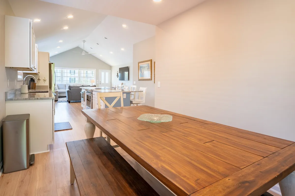 A wide view of the dining table set near the kitchen, with large windows letting in natural light and a glimpse of the adjacent sofa in the living area. The setup emphasizes the seamless transition between dining, cooking, and relaxing spaces.